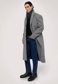 Šedý vlněný kabát s dlouhou siluetou, černý rolák, modré džíny a černé kožené boty. Šála přidává texturu a vrstvy.