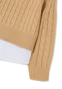 Pull en maille torsadée beige avec un ourlet et des poignets côtelés, porté par-dessus une chemise blanche à col avec une texture lisse.