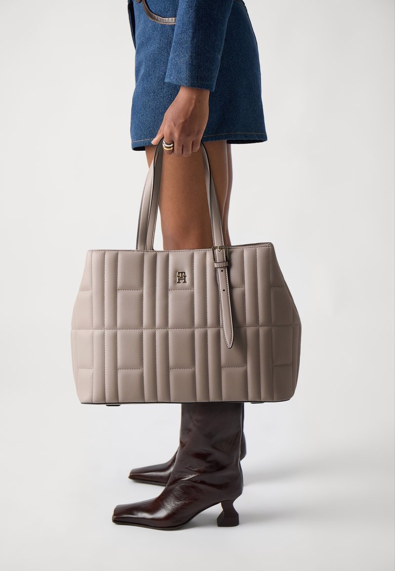 Personne portant une jupe en jean bleue et des bottes mi-mollet marron foncé, tenant un sac à main matelassé beige avec double poignée.