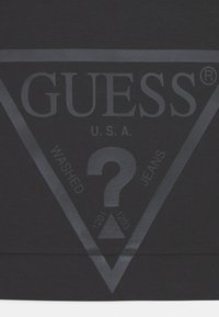 Tela oscura con un gran logo triangular que incluye la palabra "GUESS", un signo de interrogación y el texto "U.S.A. Washed Jeans" en su interior.