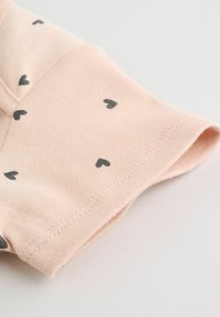 Puha rózsaszín anyag apró fekete szívmintákkal és gondosan varrott szegéllyel, egy behajtott ujj- vagy nadrágszárvégen, egyszínű világos háttéren.