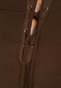 Garment in maglia marrone con scollo a V, caratterizzato da un dettaglio con lacci e occhielli dorati. Il tessuto testurizzato aggiunge profondità al design.