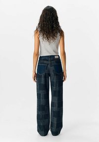 Pantalon en denim à jambes larges de couleur bleu foncé, présentant un design en patchwork avec des textures et des motifs contrastants. Poches arrière visibles.