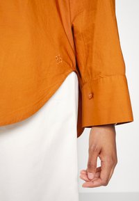 Main droite détendue à côté du corps, portant une chemise orange à manches longues avec poignets boutonnés et un pantalon blanc.