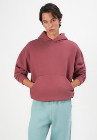 Jaunas vyras, vilkintis bordeaux spalvos hoody su rankomis prie priekinio kišenės ir šviesiai mėlynomis sportinėmis kelnėmis, priešais vientisą baltą foną.
