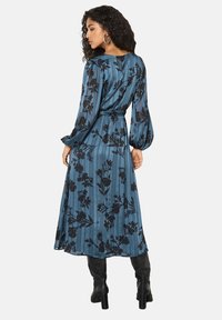 Abito a maniche lunghe color teal con motivo floreale nero e strisce verticali. Presenta una vita cintrata e una gonna fluente. Accentuato da stivali neri.