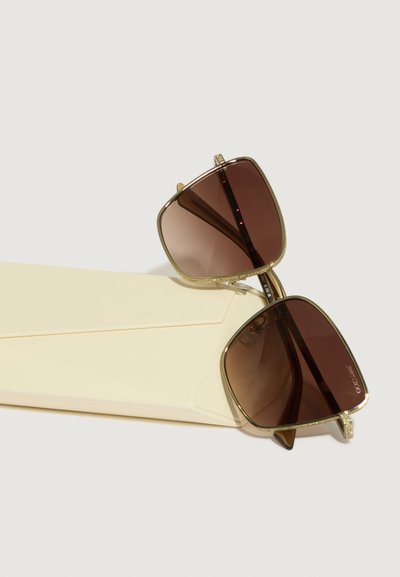 Óculos de sol quadrados castanhos com detalhes em dourado, apresentando lentes escuras e armações texturizadas, dispostos sobre uma caixa amarelo pálido.