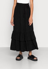 Jupe maxi noire à volants avec un motif texturé et une taille élastique. Associée à des sandales noires à lanières avec une semelle épaisse.