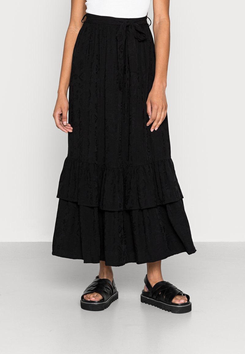 Jupe maxi noire à volants avec un motif texturé et une taille élastique. Associée à des sandales noires à lanières avec une semelle épaisse.