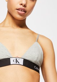 Sivi trokutasti bralette od mekanog materijala, s crnim elastičnim trakama i bijelim logotipom. Podesive tanke naramenice.