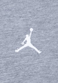 Šedá tkanina s bielym vyšívaným logom "Jumpman" v polohe vo vzduchu, ktorý drží basketbal, s textúrovaným povrchom.