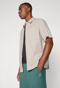 Light beige short-sleeve shirt featuring a button closure, one chest pocket, and tags, paired with green shorts and a black undershirt.