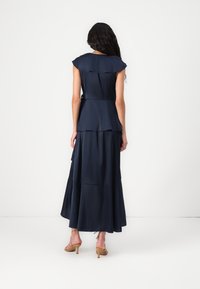 Vestido de cetim azul-marinho com design peplum, mangas curtas e esvoaçantes, e saia em camadas. Usado com saltos de tom castanho claro.