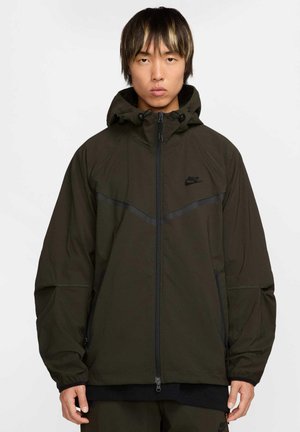 Nike takki, tumma oliivinvihreä, edessä vetoketjulukko, huppu säädettävällä nyörillä, mustat yksityiskohdat olkapäillä ja sivuilla, sileä materiaali, toiminnallinen muotoilu.