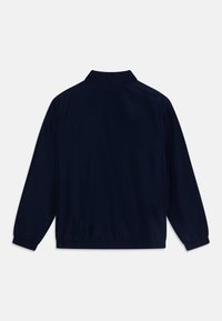 Chaqueta de manga larga azul marino con puños y dobladillo elásticos, cuello alzado, mostrada desde la parte posterior sobre un fondo blanco liso.