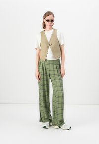 Utenermet beige vest med frontspenne, båret over en hvit T-skjorte. Grønne rutete vide bukser. Grå og grønne joggesko.