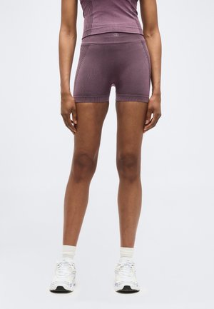 Persona che indossa pantaloncini viola aderenti, top abbinato, calzini bianchi e scarpe da ginnastica bianche, in piedi contro uno sfondo chiaro e uniforme.