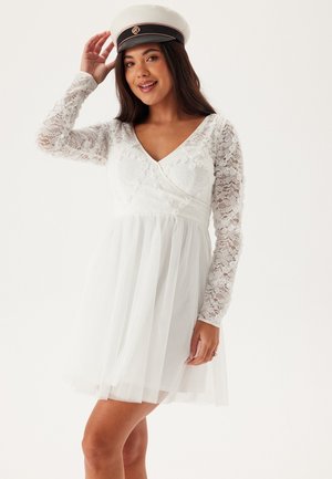 Vit klänning med spetsslangenar, figursydd överdel och flytande tyllkjol. Bärs med en dekorativ hatt som visar eleganta blommönster.