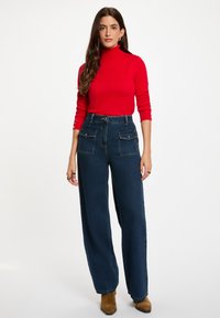 Roter Rollkragenpullover mit langen Ärmeln, kombiniert mit hoch taillierten weit geschnittenen dunkelblauen Jeans mit zwei vorderen Taschen und goldenen Beschlägen.