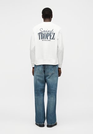 Man wearing white sweatshirt with "Saint Tropez Michael Kors" text on back and blue jeans, standing facing away on plain background.