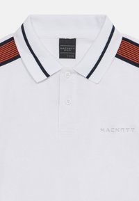 Biela polo košeľa s dvoma gombíkmi, námornícky a červené pásy na pleciach, námornícky lem na golieri a logo "Hackett" na prsiach.