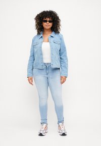 Light blue denim jacket with a fitted design, two chest pockets, paired with light blue skinny jeans and patterned sneakers. Model wearing sunglasses.