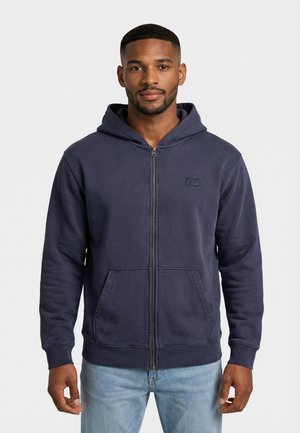 Hombre con cabello corto y barba, vistiendo una sudadera con cremallera azul marino y pantalones vaqueros azul claro, de pie contra un fondo blanco liso.