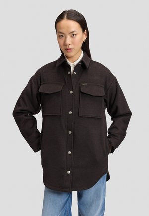 Junge Frau mit langen dunklen Haaren trägt eine schwarze Hemdjacke mit Knöpfen über einem weißen Hemd und hellblauen Jeans, steht mit den Händen in den Taschen.
