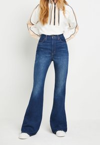 Tmavě modré vyšší zúžené džíny s jemným vyblednutím, designem s pěti kapsami a kontrastním stehem. Bílé tenisky doplňují oblečení.