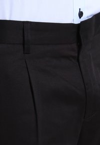 Svarta byxor med en slät, matt yta. Har en veck fram och ett bälteshål i midjan. Närbild av tyget och sömmen.