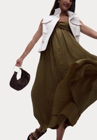 Olivgrünes, strukturiertes Maxikleid mit gerafftem Oberteil; weiße ärmellose Jacke; dunkle Handtasche mit runden Griffen; Sandalen mit Nieten.