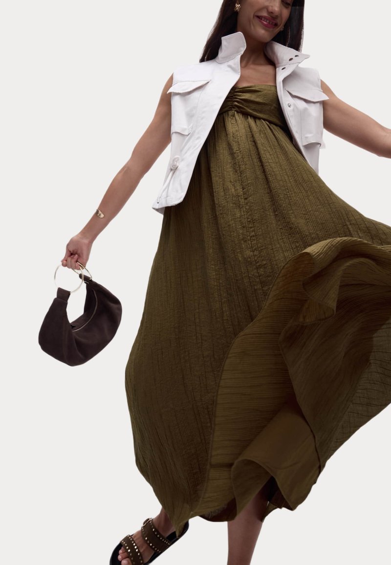 Olivgrünes, strukturiertes Maxikleid mit gerafftem Oberteil; weiße ärmellose Jacke; dunkle Handtasche mit runden Griffen; Sandalen mit Nieten.