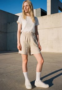 Blonde Frau steht draußen in beige-farbenen Shorts mit elastischem Bund, weißem T-Shirt, weißen Socken und weißen Turnschuhen vor einer Betonwand und blauem Himmel.