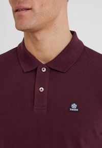 Burgundy polo shirt made of textured fabric, featuring a collar, three buttons, and a black logo patch on the chest.