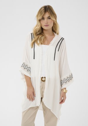 Donna bionda con pantaloni beige, top bianco e kimono bianco ampio con ricamo nero e frange sulle maniche e sui bordi.