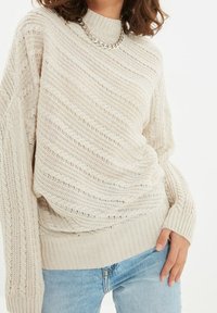 Pull en maille crème avec un motif diagonal texturé, un ourlet côtelé et un col rond. Assorti à un jean bleu clair. Accent chaîne argentée visible.