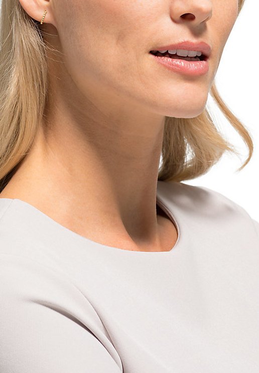 Gul hoopörhänge som bärs i vänster öra, med en texturerad design. Subjektet har på sig en ljusgrå blus med rundad halsringning.