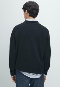 Czarny dzianinowy sweter z kołnierzykiem, ściągaczami na manKietach i luźnym krojem, noszony na białą koszulę, widziany od tyłu.