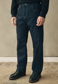 Jeans de mezclilla azul oscuro con un corte recto, costuras visibles y bolsillos frontales, combinados con zapatos negros de tipo slip-on.