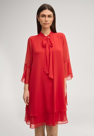 Frau trägt ein knielanges rotes Chiffonkleid mit transparenten Ärmeln und einer gebundenen Schleife am Ausschnitt, steht vor einem einfachen Hintergrund.