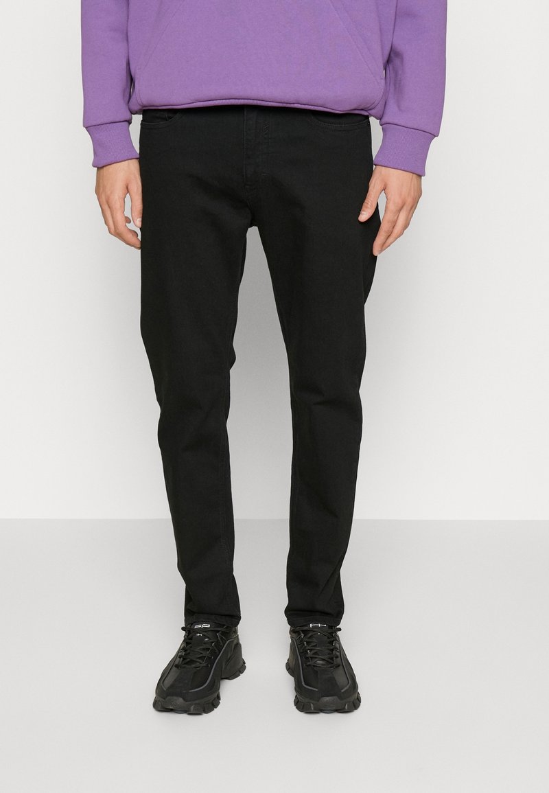 Jean en denim noir avec une coupe slim, un devant plat et des poches latérales, associé à un sweat-shirt violet et des baskets de sport noires.