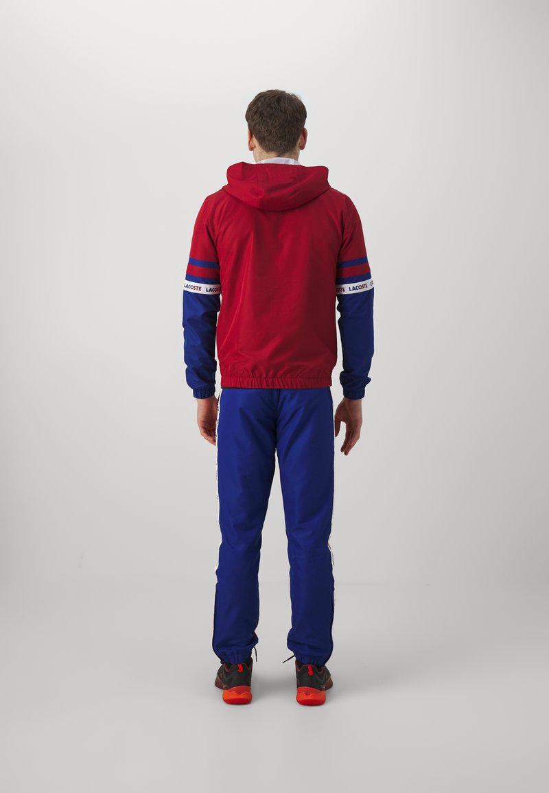 Rood en blauw sporttrackuit met capuchon, voorzien van elastische manchetten en tailleband, geïntensiveerd met branding op de mouwen. Zwarte schoenen met rode zolen.