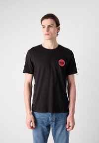 Black cotton t-shirt with short sleeves, round neckline, and red circular "HUGO" logo on the left chest. Worn with blue denim jeans.