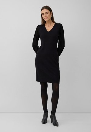 Woman wearing a black knee-length dress with sheer long sleeves, black tights, and black heeled ankle boots against a plain gray background.