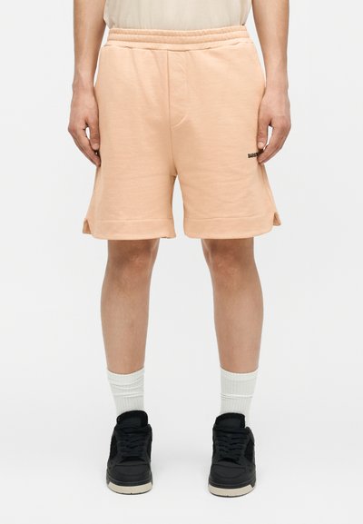 Άντρας που φοράει ανοιχτόχρωμα ροδάκινα ελαστικά σορτς με μικρό μαύρο λογότυπο στο αριστερό μηρό, λευκές κάλτσες και μαύρα αθλητικά παπούτσια, όρθιος.
