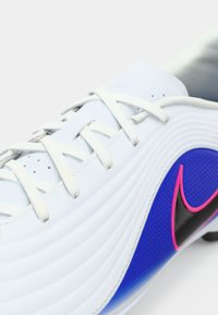 Λευκό αθλητικό παπούτσι με λευκές κορδόνες και μαύρο λογότυπο Nike περιγραμμένο σε ροζ, σε μπλε υφαντό φόντο.