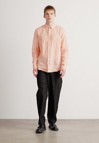 Lys rosa lin skjorte med knapper og lange ermer kombinert med svarte bukser. Modellen står med uttrykksløs ansikt og svarte sko.