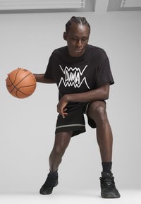 T-shirt noir avec le logo "PUMA" blanc, shorts noirs avec une bande grise, tenant un ballon de basket, portant des baskets noires. Cadre intérieur avec un fond minimal.
