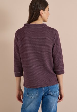 Femme vue de dos portant un haut à manches longues rayé violet et blanc et un jean bleu sur fond beige.