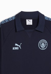 Polo blu marino con colletto nero, dettagli azzurri e motivo grafico sulle maniche. Presenta il logo 'KING' e l'emblema del Manchester City.